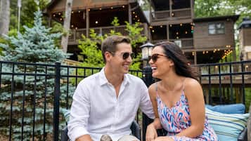 Un couple est assis sur un banc, avec une terrasse en bois et des guirlandes lumineuses en arrière-plan.