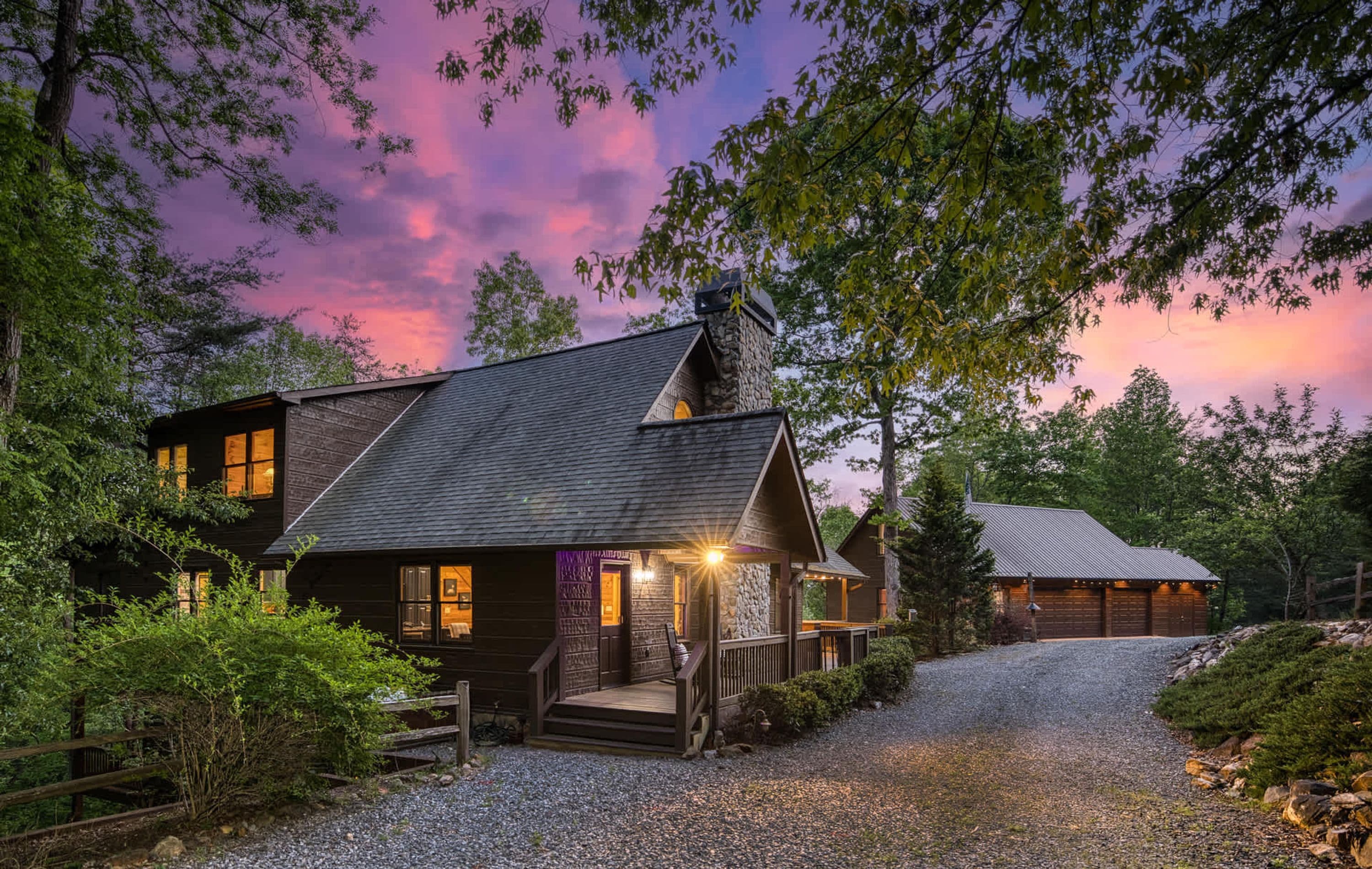 A rustic cabin with a stone chimney, wooden exterior, and a gravel driveway leading to a wooden deck.