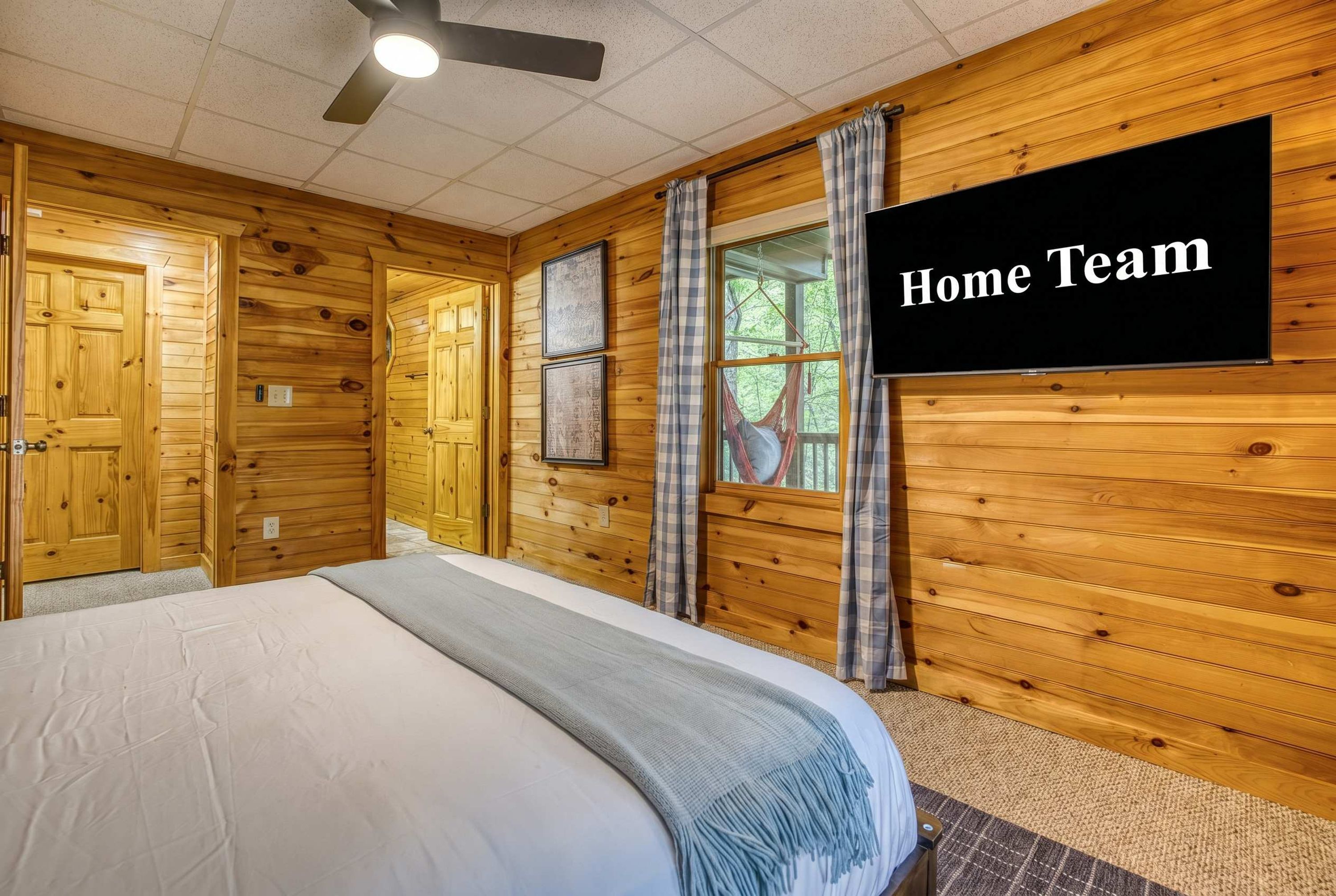A room with wooden walls, a bed, a ceiling fan, a window with curtains, and a flat-screen TV.
