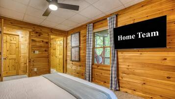 Une pièce aux murs en bois, avec un lit, un ventilateur de plafond, une fenêtre avec des rideaux et un téléviseur à écran plat.