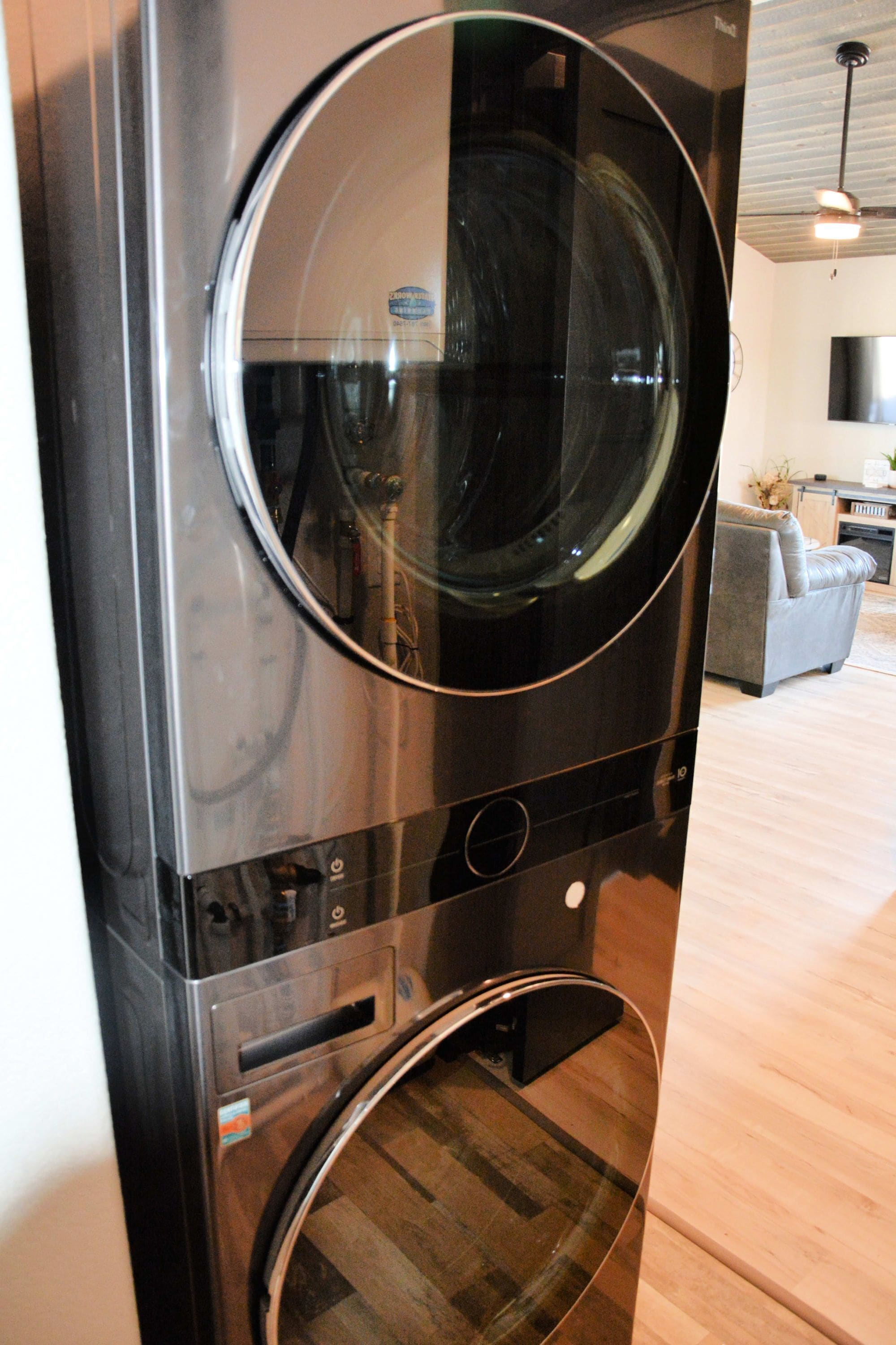 A stacked washer and dryer unit in a modern kitchen with a wooden floor and a grey sofa in the background.