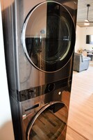 A stacked washer and dryer unit in a modern kitchen with a wooden floor and a grey sofa in the background.
