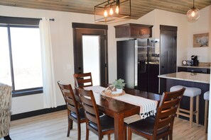 Un comedor con una mesa de madera, sillas y una zona de cocina con refrigerador y cafetera.