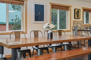 Un comedor con mesa de madera, sillas metálicas y un banco. Hay ventanas con persianas, un cuadro en la pared y un salpicadero de piedra en la zona de la cocina.