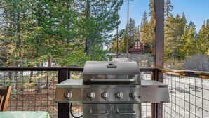 Una parrilla con cuatro quemadores, una mesa con mantel a cuadros y vista a una cabaña en un bosque.