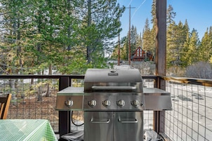Una parrilla con cuatro quemadores, una mesa con mantel a cuadros y vista a una cabaña en un bosque.