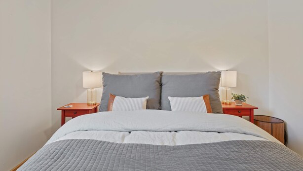 Una cama bien hecha con colcha gris, dos mesitas de noche con lámparas y un cabecero grande en tono gris.