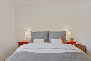 Una cama bien hecha con colcha gris, dos mesitas de noche con lámparas y un cabecero grande en tono gris.