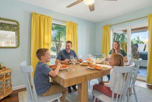 Eine Familie isst an einem Tisch, im Hintergrund gelbe Vorhänge, ein Deckenventilator und Blick auf Grünfläche.