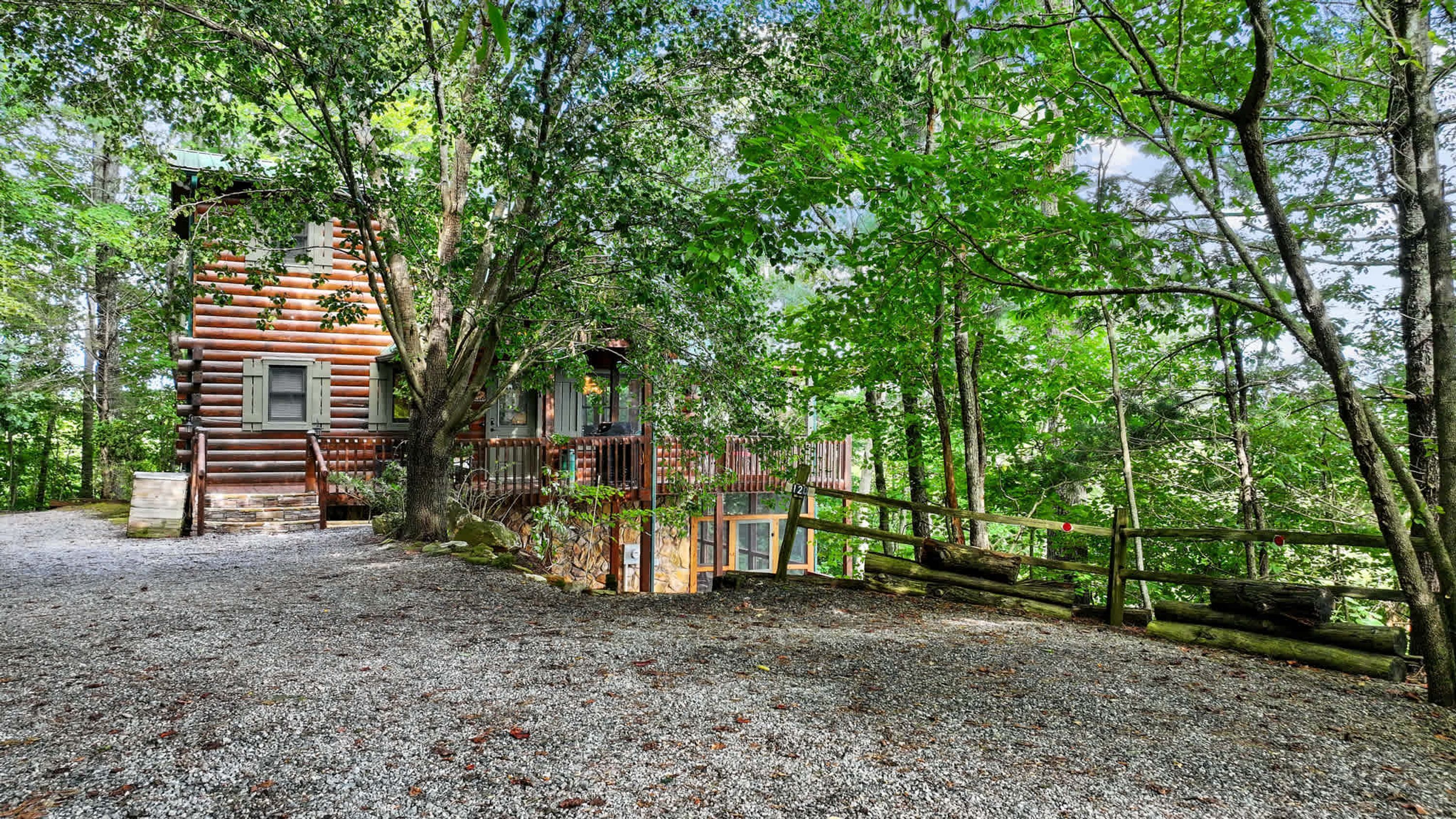 A rustic log cabin with a gravel driveway, surrounded by dense forest.
