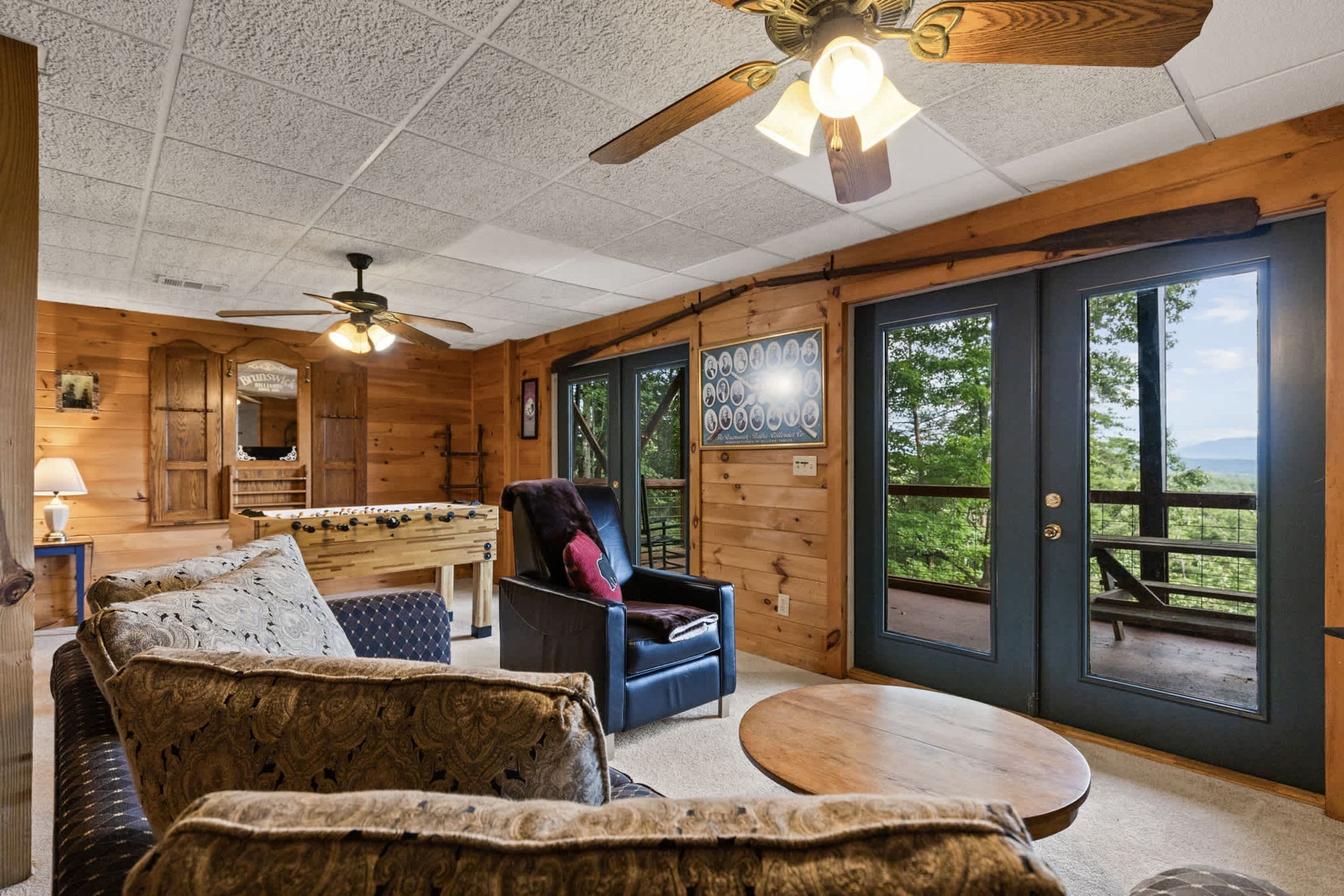 A cozy room with a ceiling fan, a wooden table, a sofa, and a chair. There are large windows with a view of greenery outside.