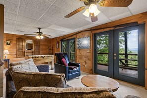 A cozy room with a ceiling fan, a wooden table, a sofa, and a chair. There are large windows with a view of greenery outside. - Cabin Sweet Cabin Hot Tub Game Room Dog Friendly (Morganton)