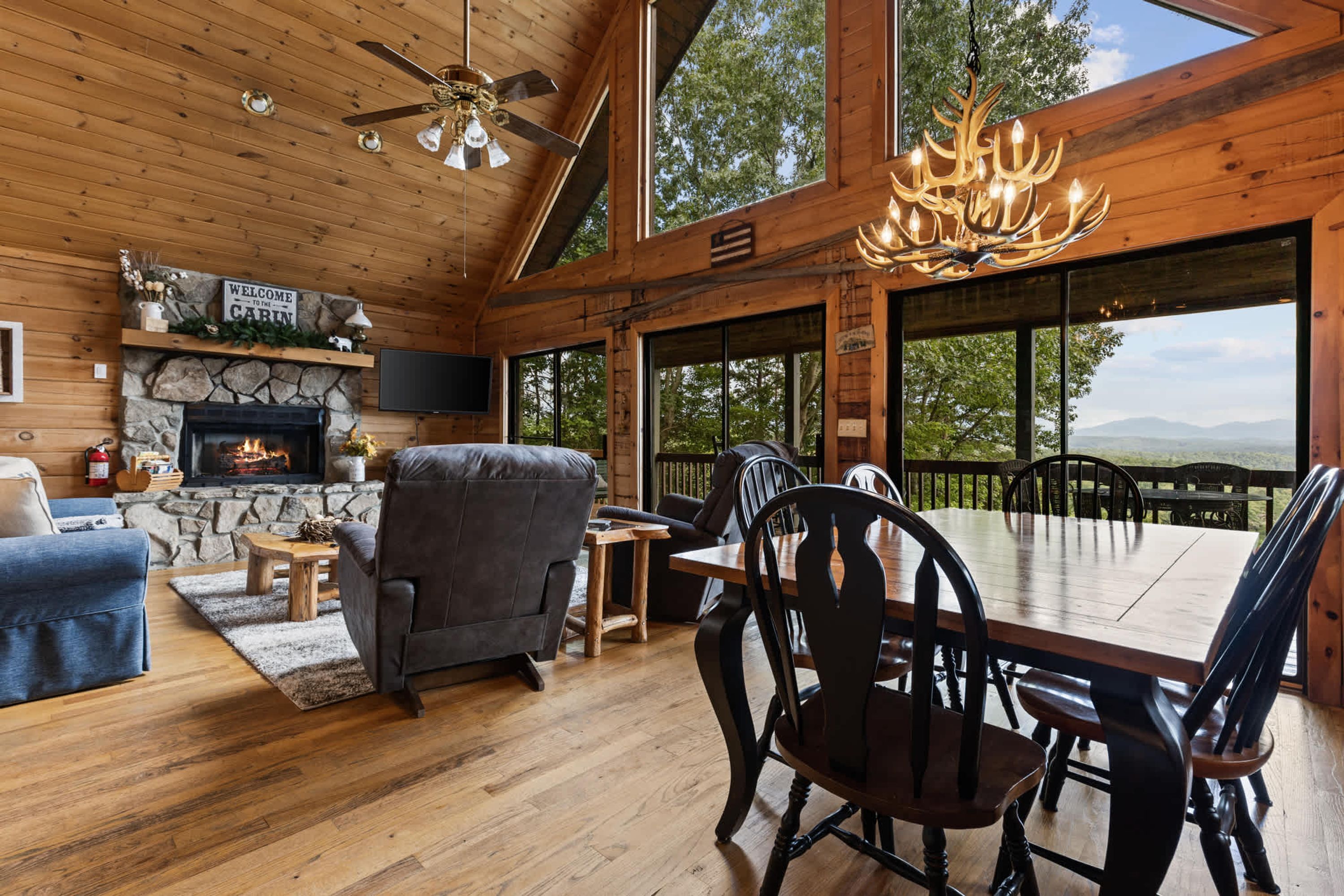 A cabin interior with a stone fireplace, a ceiling fan, a dining table with chairs, and a large window offering a view of the outdoors.