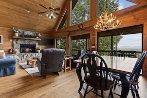 A cabin interior with a stone fireplace, a ceiling fan, a dining table with chairs, and a large window offering a view of the outdoors.