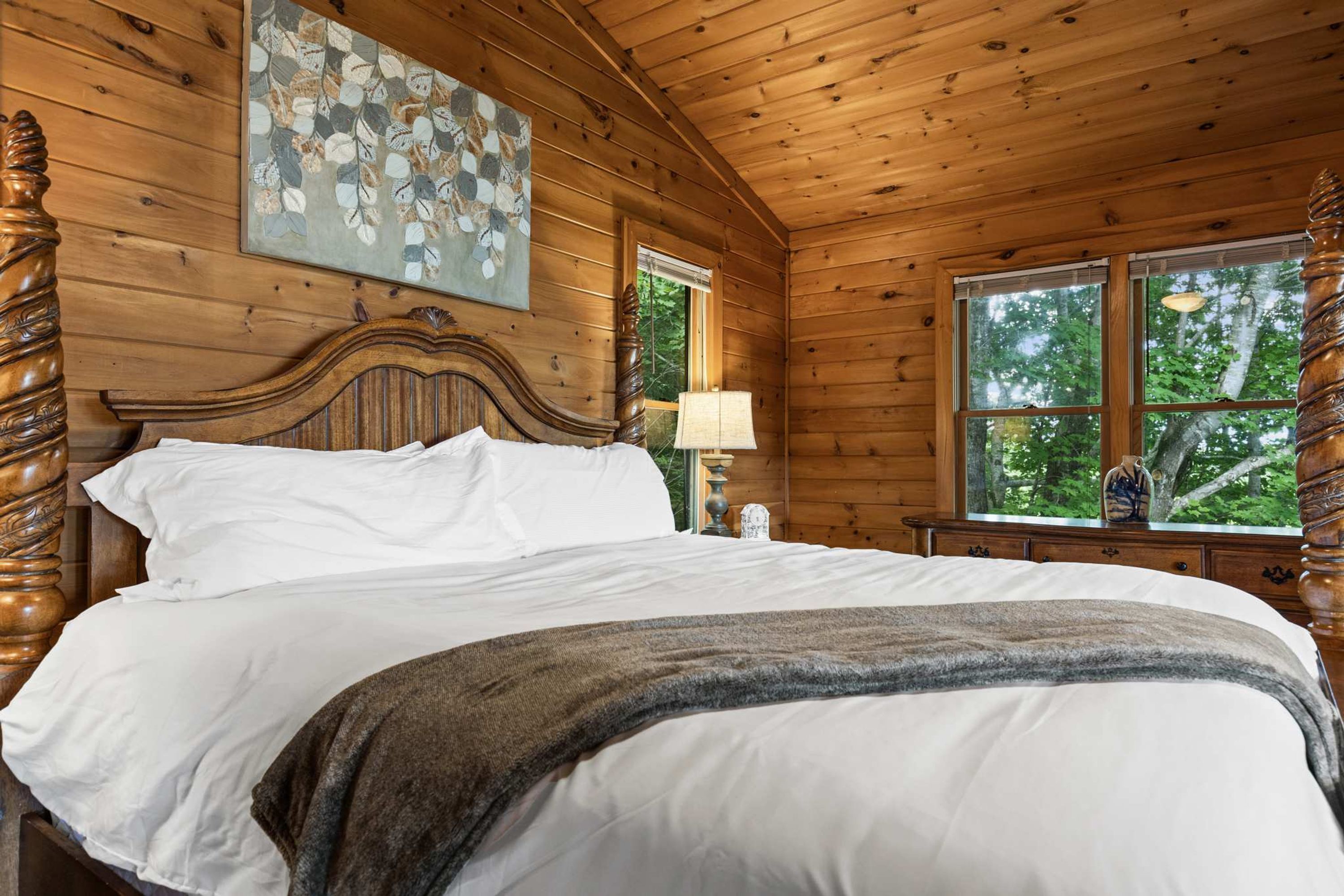 A wooden cabin bedroom with a large bed, a painting above the bed, and a window with a view of trees.