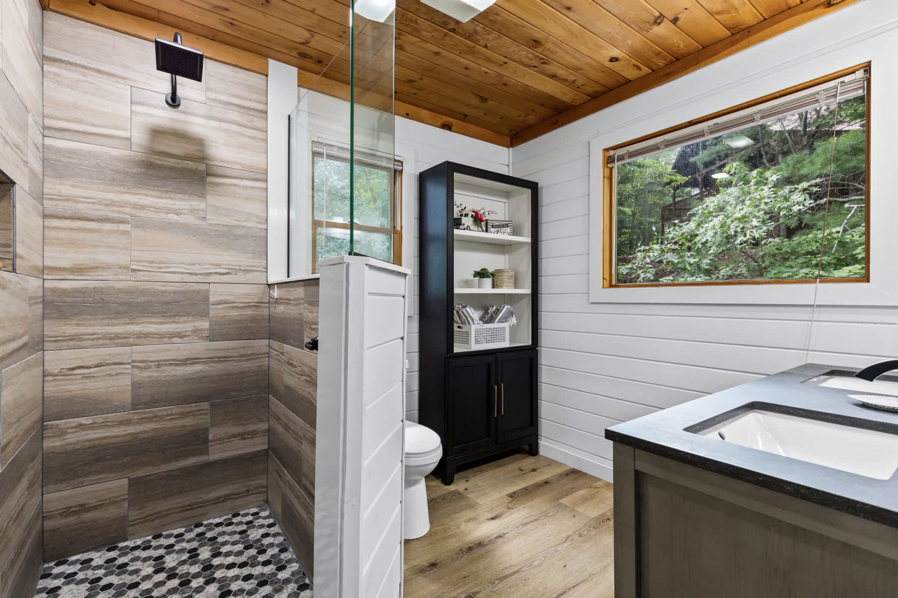 A bathroom with a shower, toilet, and sink, wooden flooring, and a window with a view of greenery.