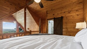 Ein Schlafzimmer mit einem großen Bett, Holzbalkendecke, Blick durch ein Fenster und einer Tür.