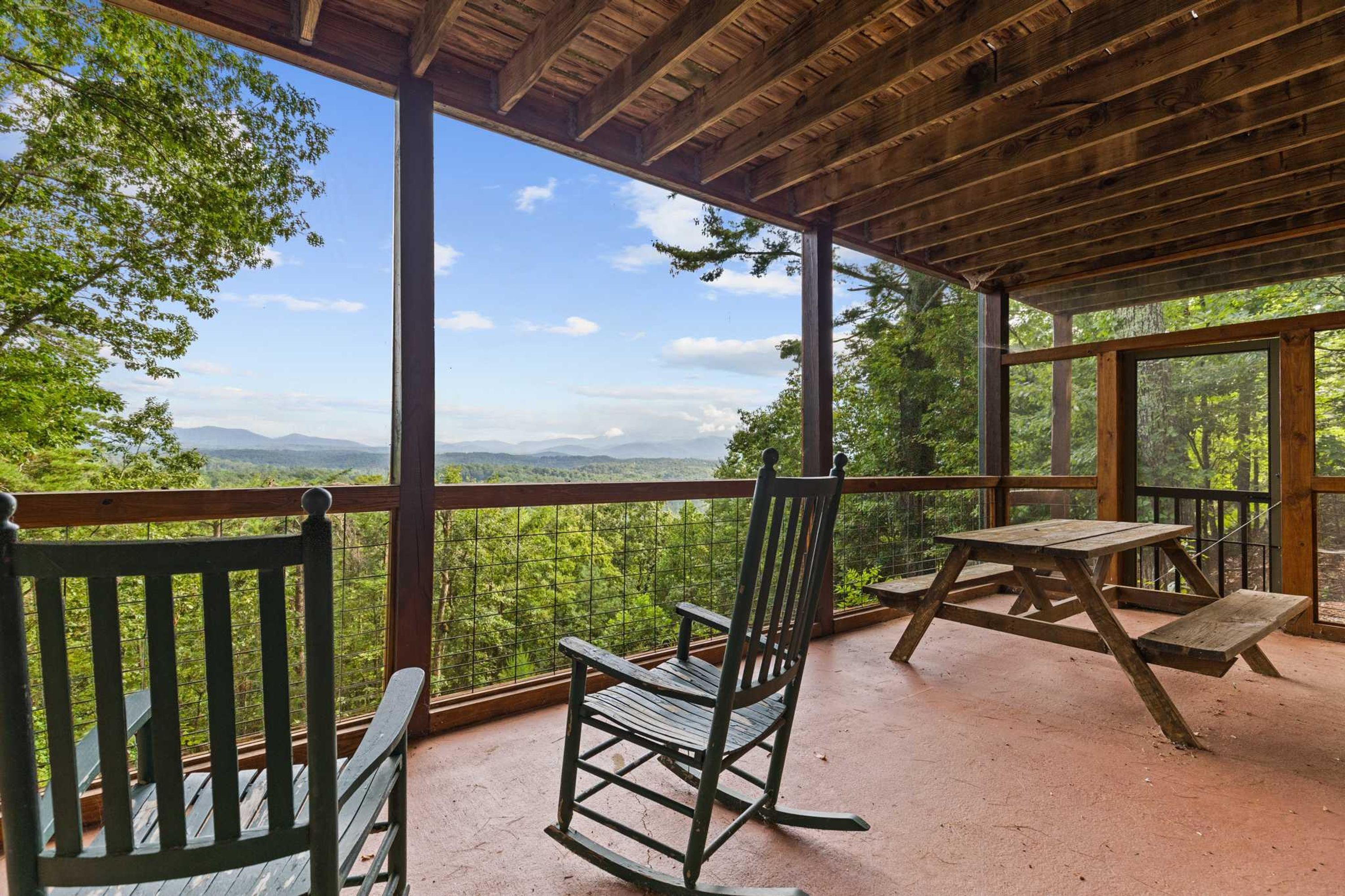 Terraza de madera con vistas a un bosque y montañas. Hay dos mecedoras y una mesa de picnic.