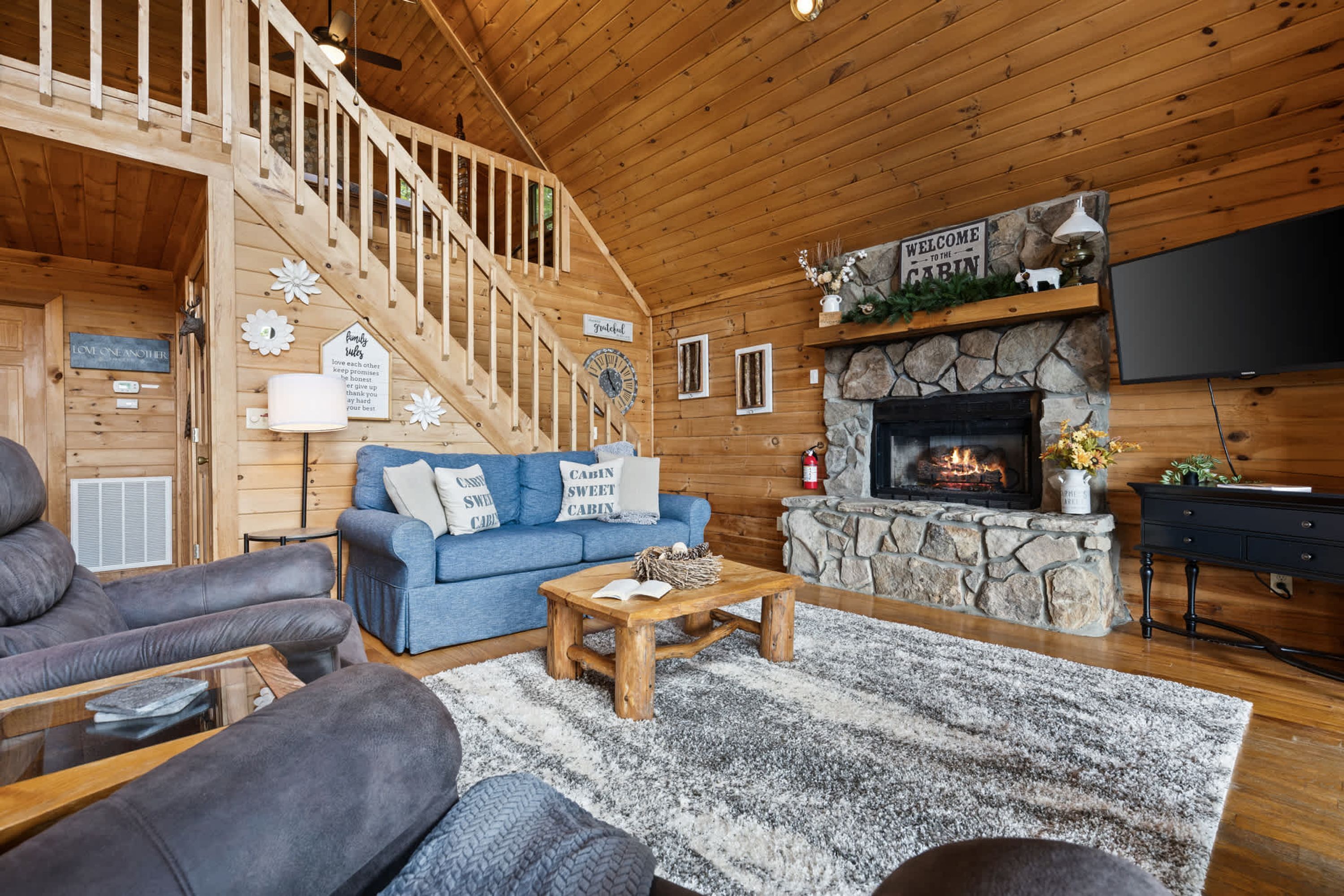 A cozy cabin interior with a stone fireplace, a blue sofa, a wooden staircase, and a television mounted above the fireplace.