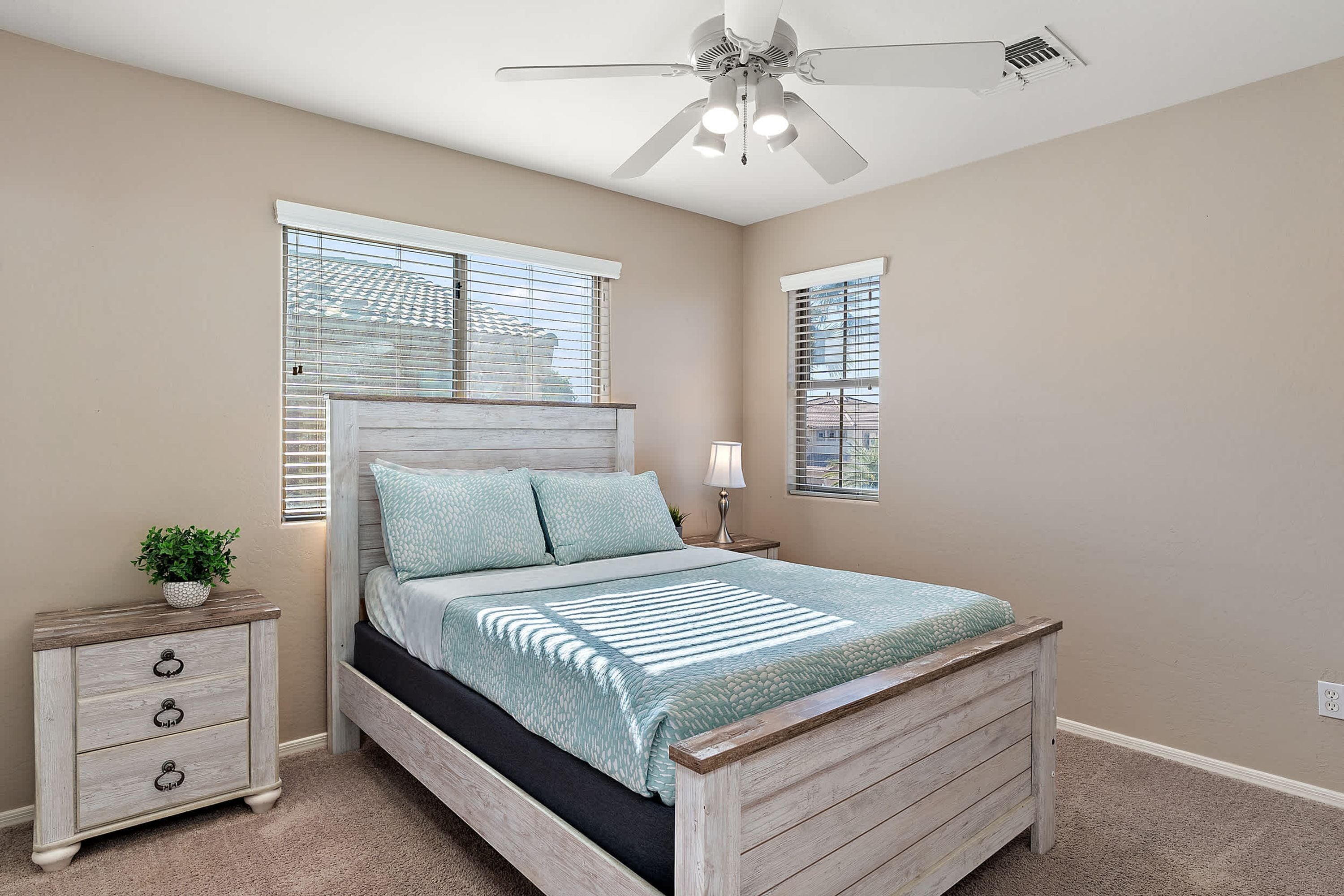 A bedroom with a bed, a nightstand, a lamp, a ceiling fan, and windows with blinds.