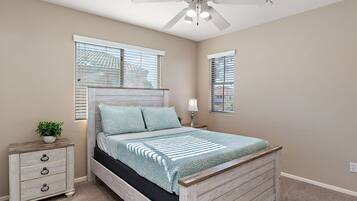 A bedroom with a bed, a nightstand, a lamp, a ceiling fan, and windows with blinds.