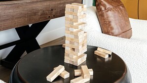 Ein Wohnzimmer mit einem hölzernen Jenga-Turm auf einem schwarzen Couchtisch, einem weißen Sofa, einem braunen Lederkissen und einem Fernseher auf einem Holzständer.