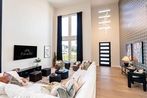 A modern living room with a sofa, coffee table, and TV. There is a large window with curtains, a door, and a wall with a geometric pattern. - Old Town Estate Pool Theater Game Room (Scottsdale)