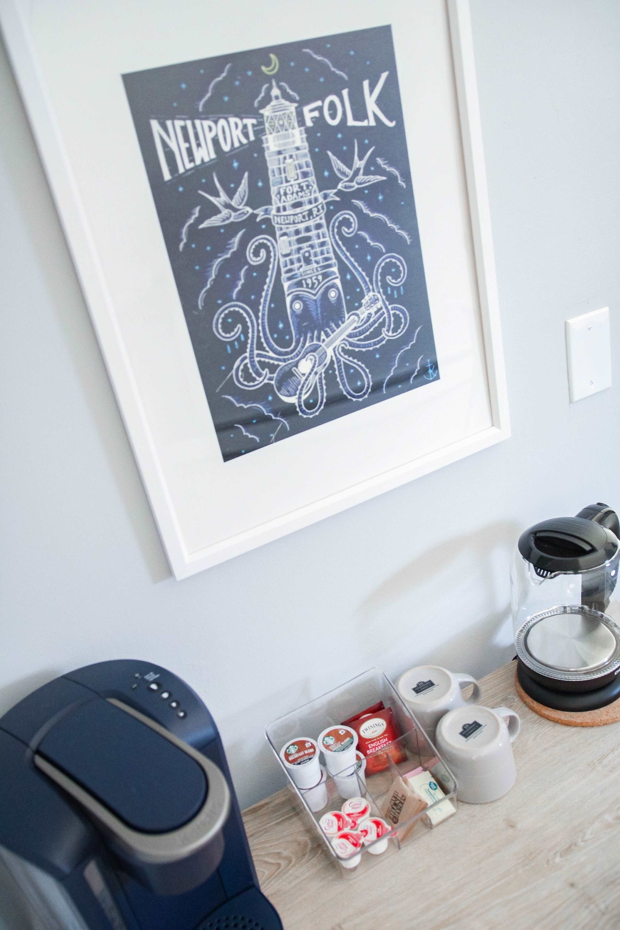 A framed artwork of a lighthouse with the text 'Newport Folk' and 'Fort Adams' on a wall. Below, there's a coffee maker, a kettle, and a tray with tea bags and cups.