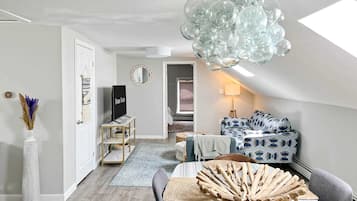 A modern loft bedroom with a sloped ceiling, a large hanging light fixture, a bed with patterned bedding, and a view into the adjacent living area.