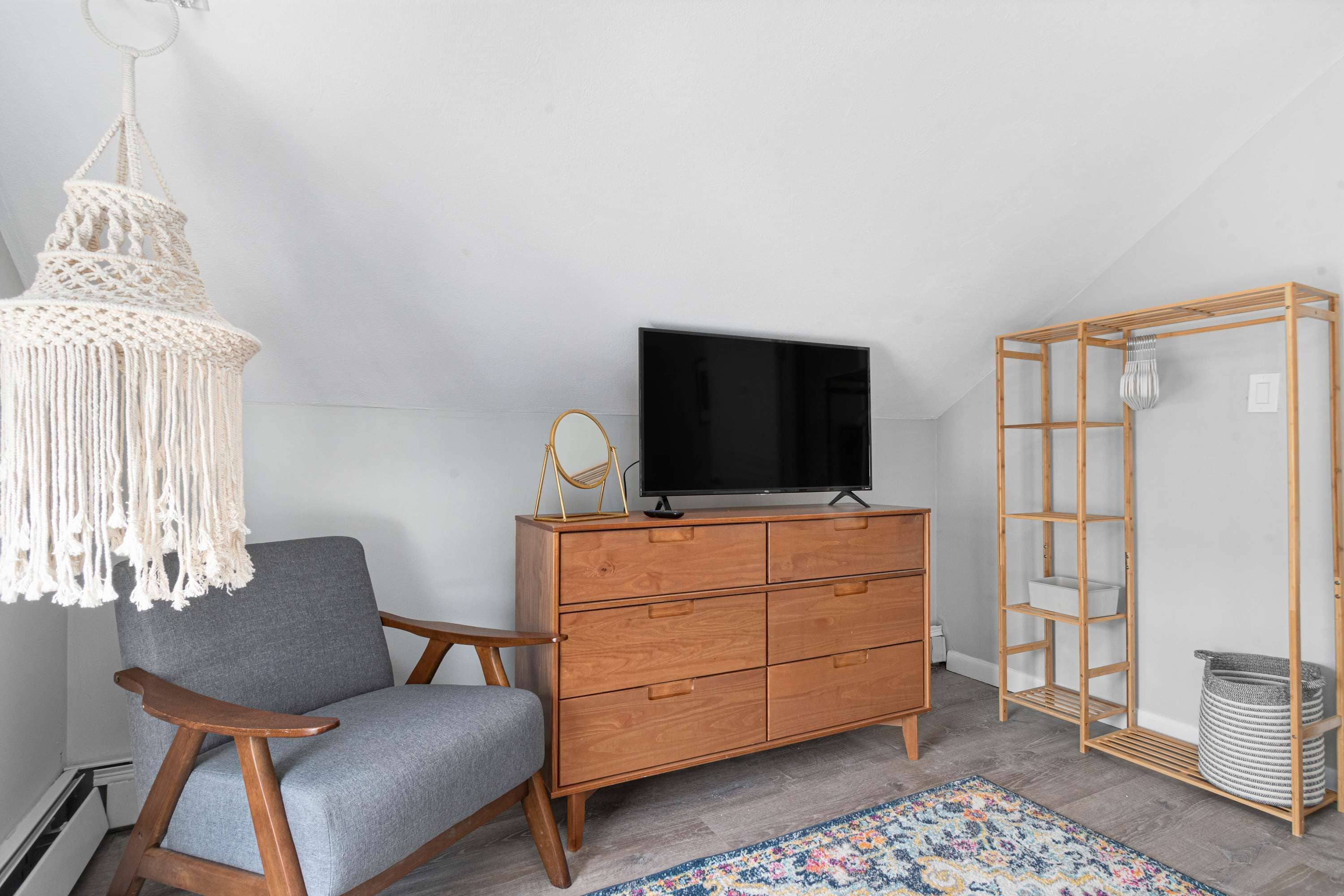 A room with a grey armchair, wooden dresser, TV, macrame light, and a woven shelf unit.