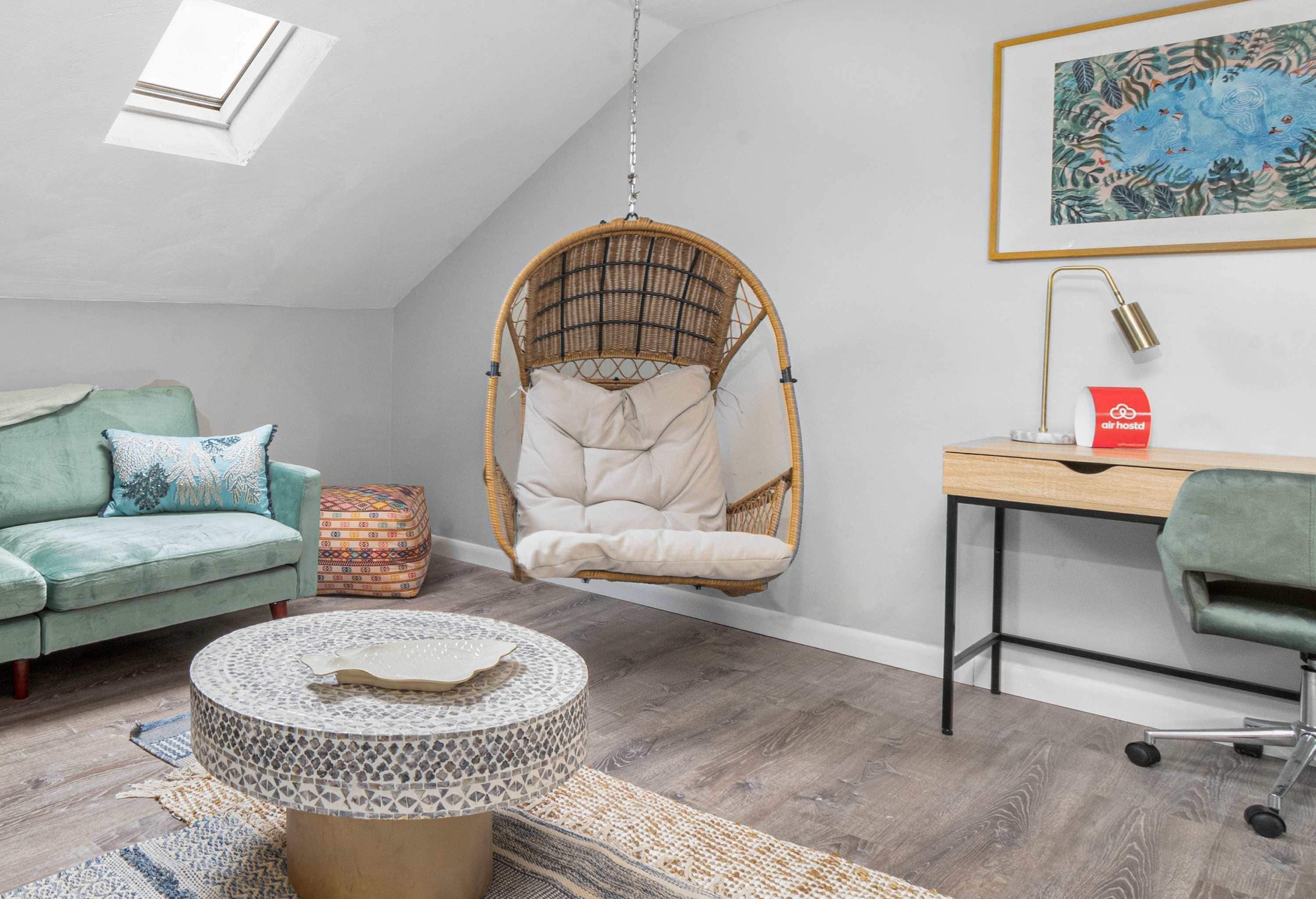 A cozy room with a hanging chair, a desk, a cushioned bench, and a round table with a patterned cover.