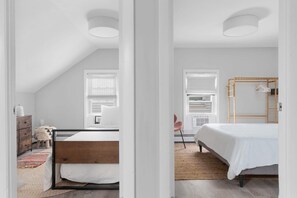 A bedroom with a sloped ceiling, a wooden bed frame, a white bedspread, a wooden wardrobe, and a small stool with a patterned cushion.