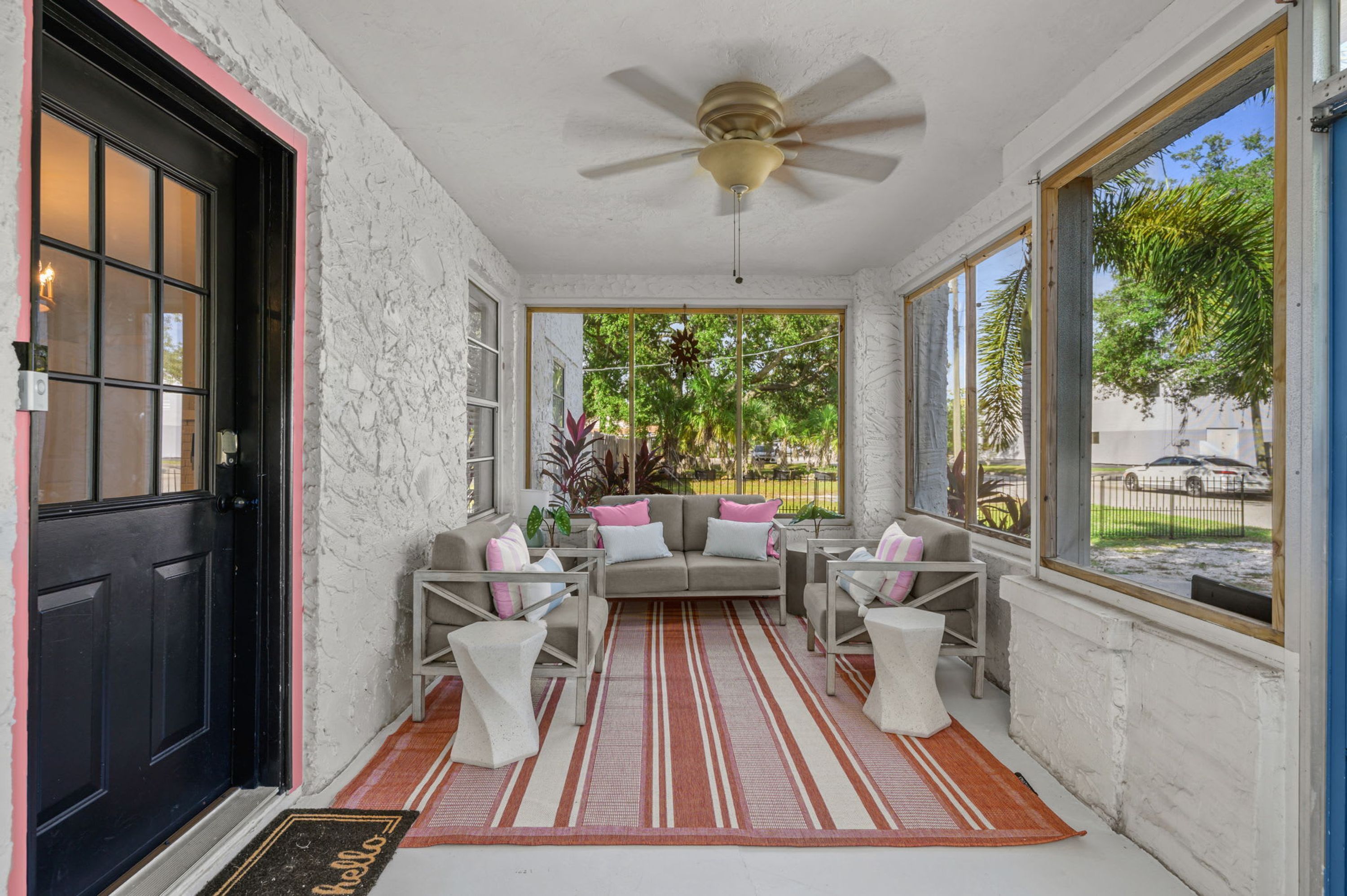 Eine sonnendurchflutete Veranda mit Deckenventilator, einem grauen Sofa mit rosafarbenen Kissen und einem Streifen Teppich.