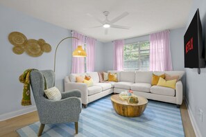 Ein Wohnzimmer mit grauem Sessel, einem weißen Ecksofa, einem runden Couchtisch aus Holz und einem Deckenventilator.
