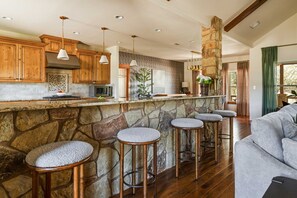 A kitchen with wooden cabinets, a stone island with bar stools, and a stone fireplace. - Stillstone Ranch A Wellness Retreat Sleeps 32 (Driftwood)