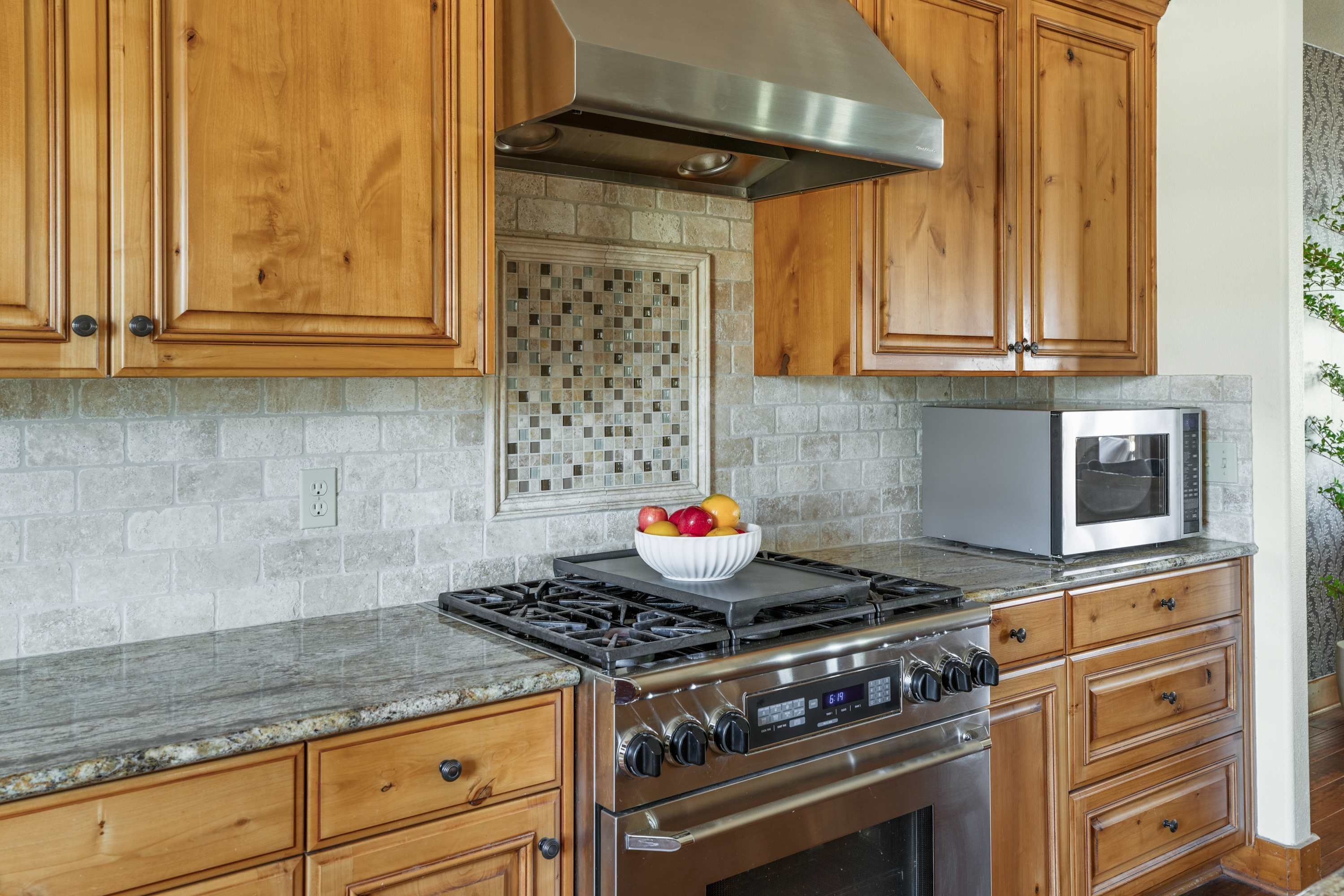 A kitchen with wooden cabinets, a stainless steel range hood, a microwave, and a bowl of fruit on the counter.