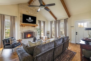 A spacious living room with a stone fireplace, a ceiling fan, and a flat-screen TV mounted on the wall. - Stillstone Ranch A Wellness Retreat Sleeps 32 (Driftwood)