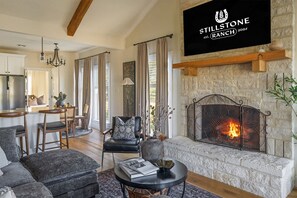 A modern living room with a stone fireplace, a dining area, and a kitchen with a stainless steel refrigerator. - Stillstone Ranch A Wellness Retreat Sleeps 32 (Driftwood)