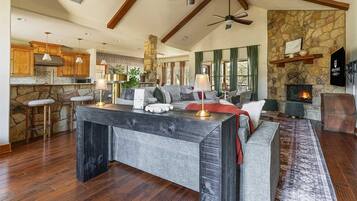 A spacious living area with a stone fireplace, a large island table, and a high ceiling with exposed beams.