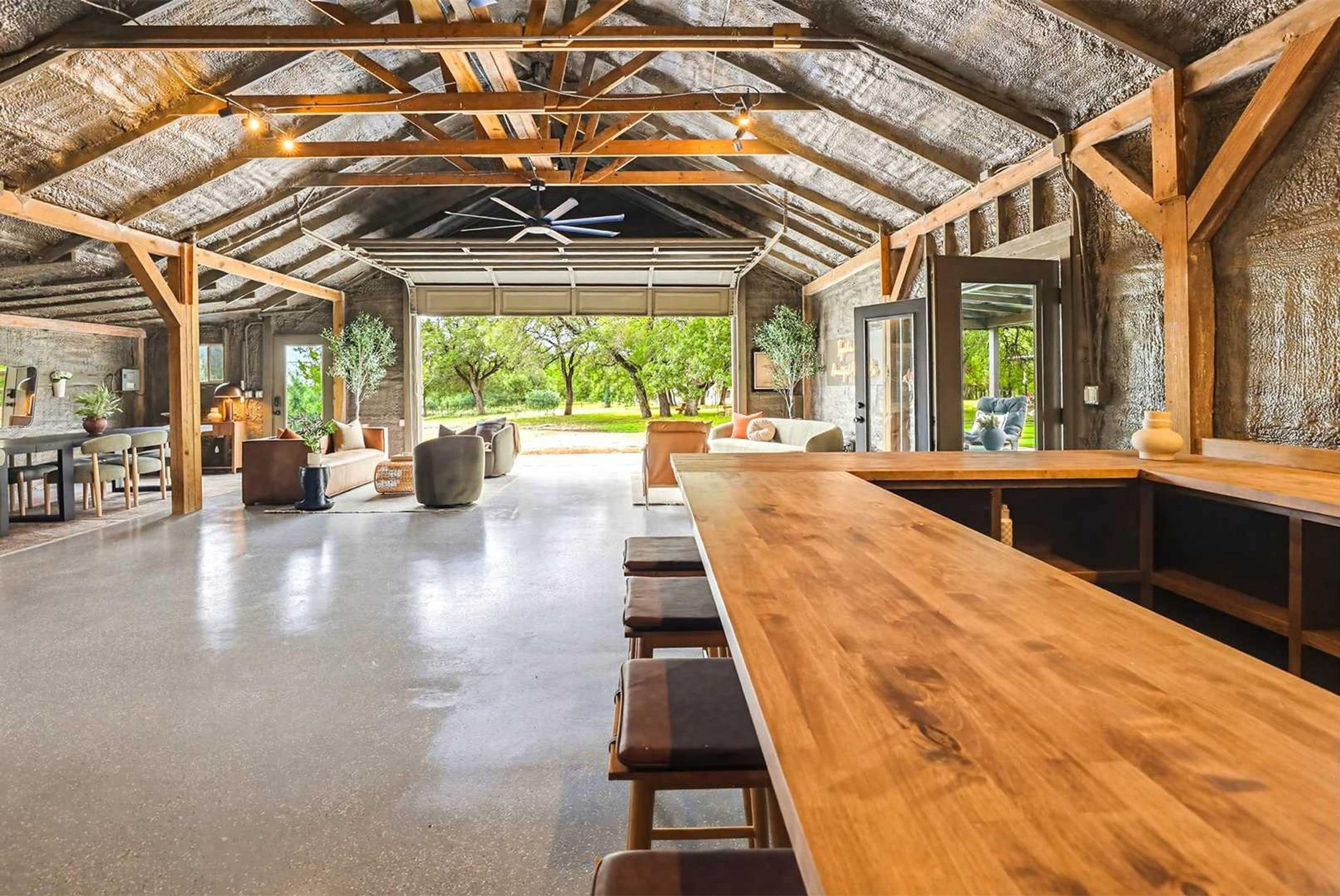 Un amplio espacio cubierto para comer con una larga mesa de madera y bancos, rodeado de muros de piedra y ventanas grandes.