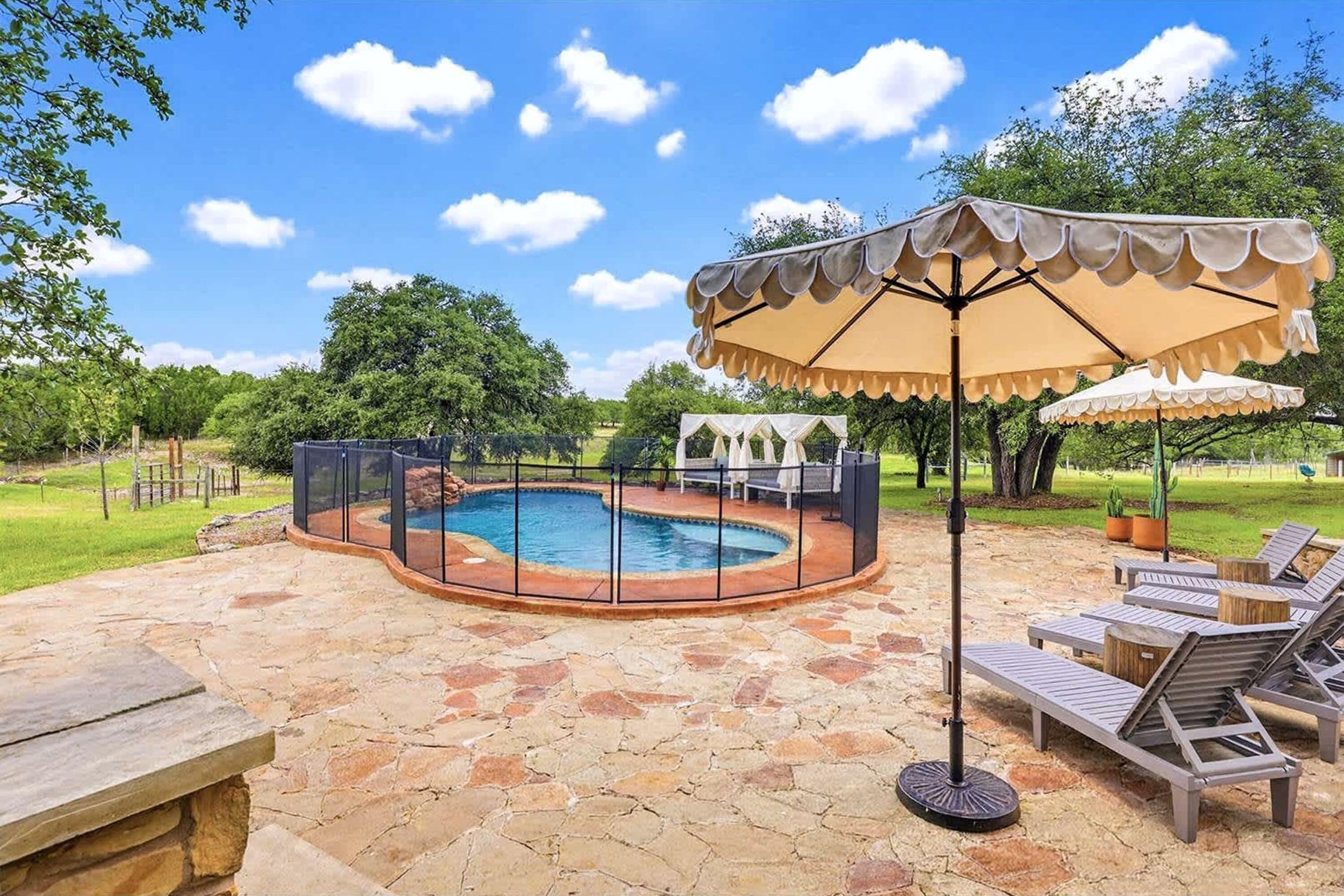 An outdoor swimming pool with a stone patio, surrounded by a fence, and shaded by a large umbrella.