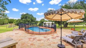 An outdoor swimming pool with a stone patio, surrounded by a fence, and shaded by a large umbrella.