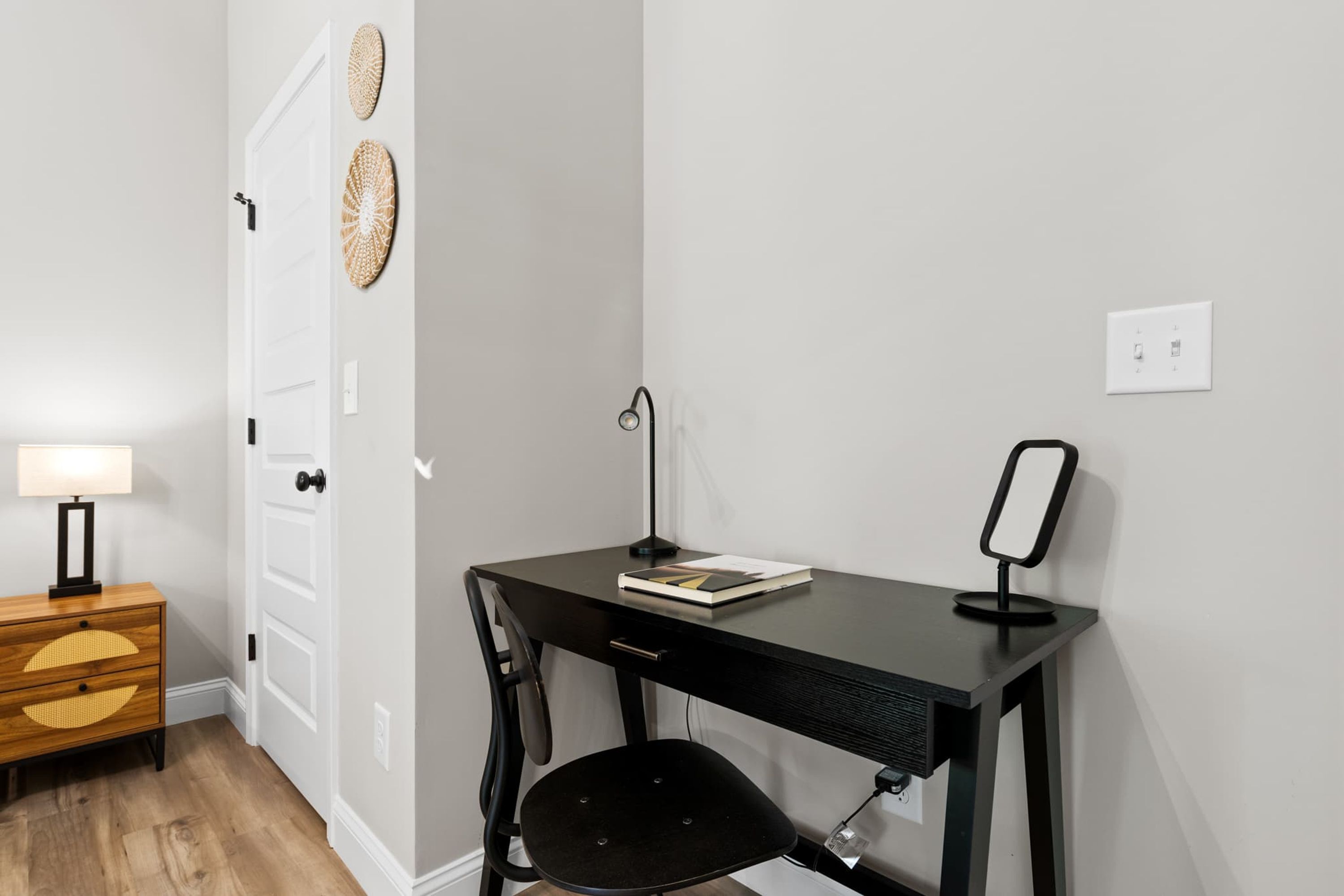 A compact workspace with a desk, chair, and a small bedside table with a lamp.