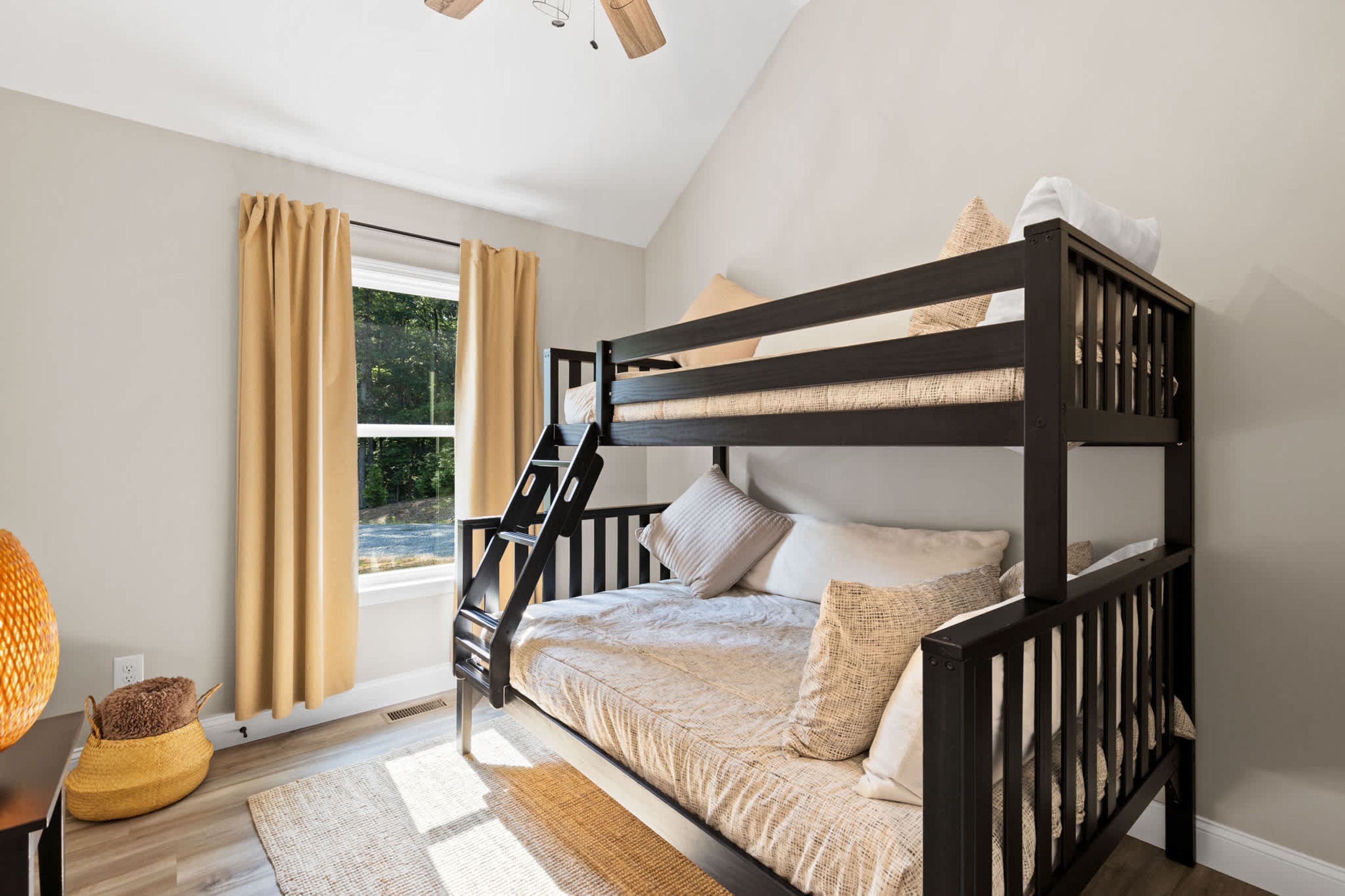 Une chambre avec un lit superposé, une fenêtre, des rideaux, un panier en osier et un plancher en bois.