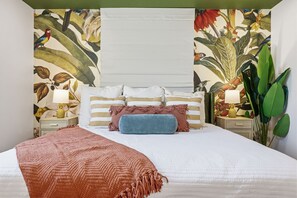 A bedroom with a bed, pillows, a blanket, a nightstand with a lamp, and a wall with a tropical wallpaper. - Epic Backyard - Pool - Hot Tub - Outdoor Theatre (Bradenton)