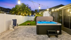 A hot tub in a backyard with a white fence, palm trees, and string lights. - Breezy Livin - Speakeasy - Pool - Spa - Game Room (Bradenton)