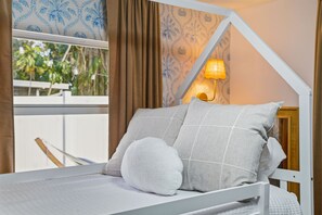 Ein Bett mit weißem Gestell, grauer Bettwäsche und einem weißen Kissen. Eine Wandlampe mit gelber Schirm.