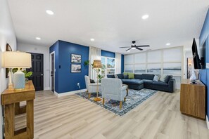A modern living room with a blue accent wall, a large window, and a ceiling fan. - Breezy Livin - Speakeasy - Pool - Spa - Game Room (Bradenton)