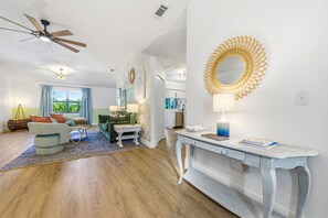Un salon moderne avec un ventilateur de plafond, un coin repas avec une table et des chaises, et une cuisine visible à travers une porte ouverte.