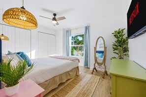 Une chambre à coucher comprenant un lit, un ventilateur de plafond, une fenêtre avec des rideaux, un téléviseur fixé au mur et une armoire verte.